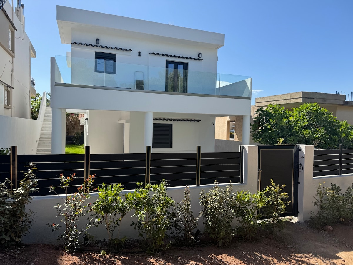 A modern two-story villa is displayed, featuring a glass railing balcony on the upper level. The white exterior contrasts with the green garden and shrubbery in the foreground. A black fence encloses the property, providing a sense of privacy and security.