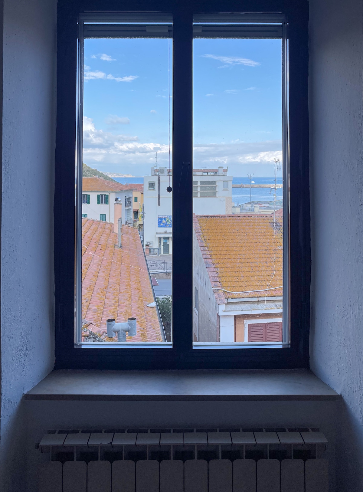 A window frames a view of rooftops overlooking a seaside landscape. The bright blue sky is partially filled with clouds, while the distant sea can be seen glistening under the sunlight. Warm tones of terracotta rooftops contrast with the coolness of the view.