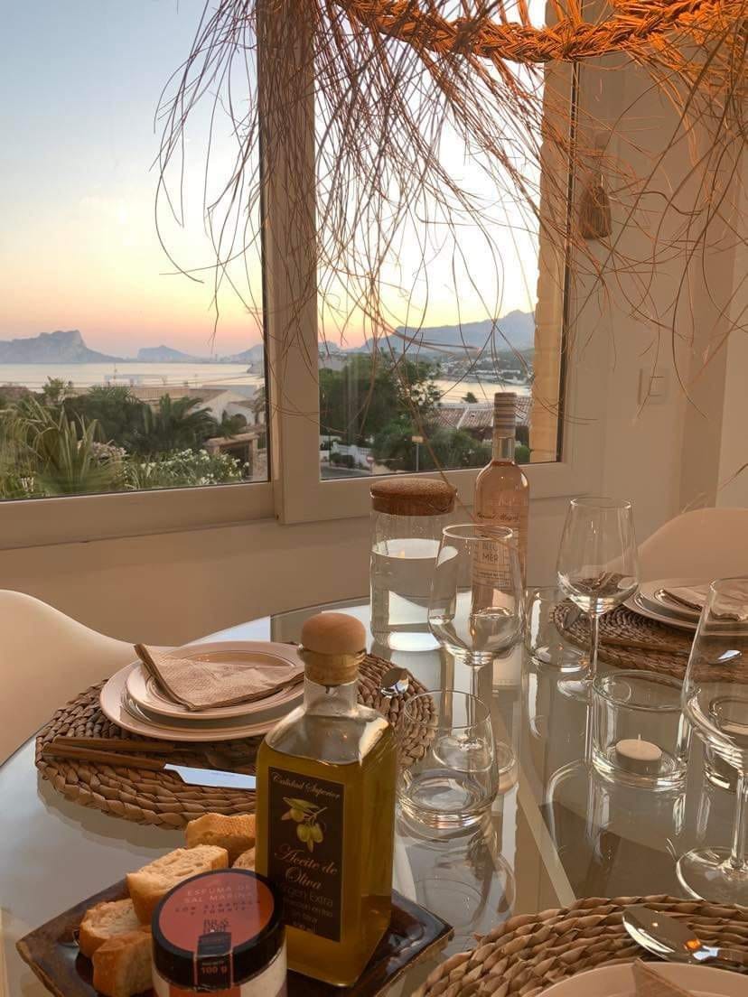 A dining table is set elegantly, featuring glassware and woven placemats. An olive oil bottle and small jars are arranged among the plates. The backdrop displays a serene sunset over the coastline, with soft hues illuminating the landscape through large windows.