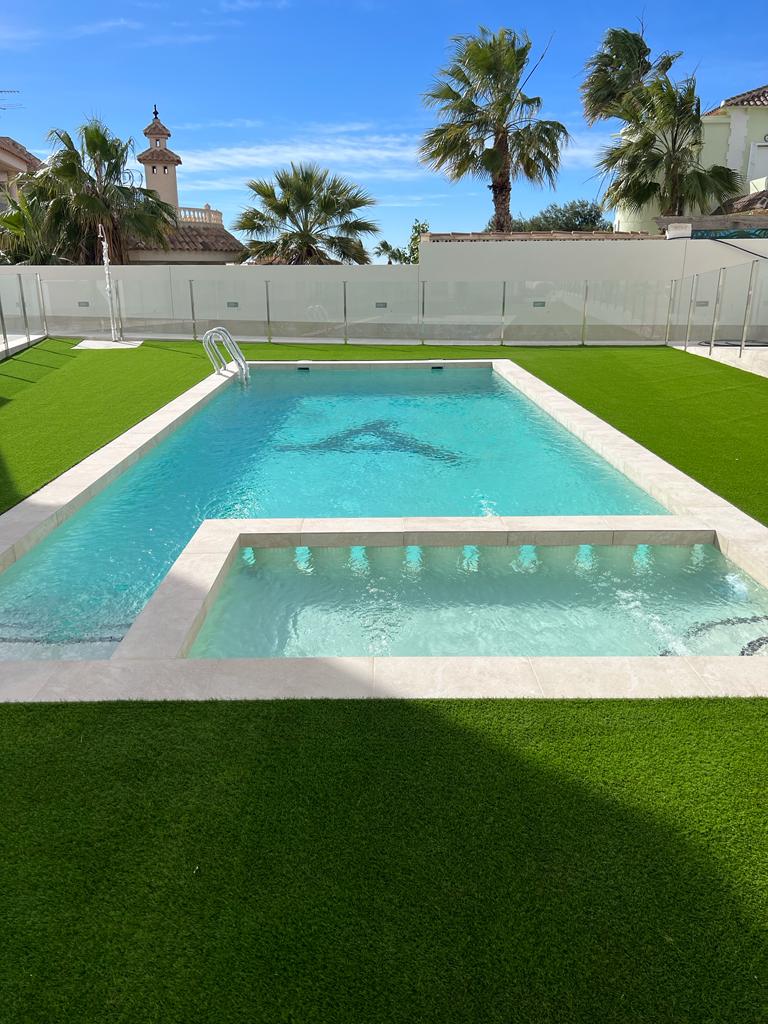 A private pool is surrounded by lush artificial grass and glass fencing, providing an open and inviting space. The pool features clear blue water and a connected hot tub. Palm trees frame the area under a bright blue sky, enhancing the serene environment.