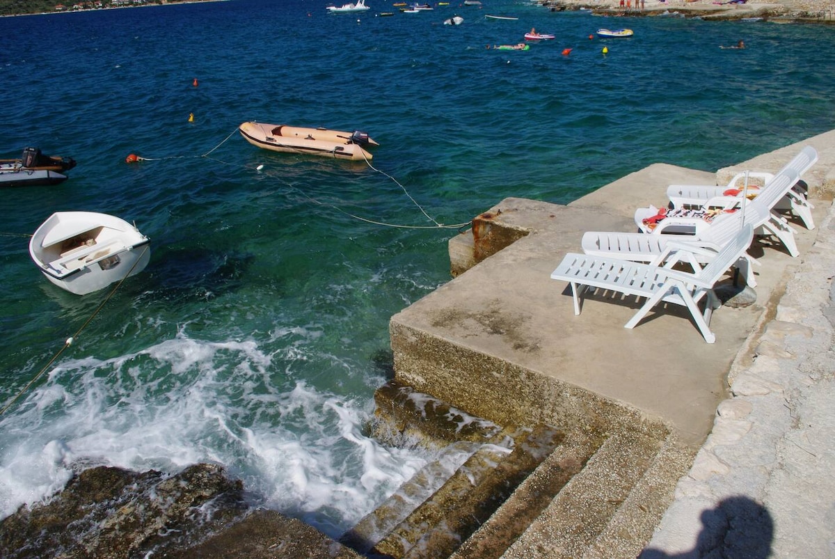 A sun-soaked waterfront area features a stone platform with steps leading down to the clear blue water. Two white lounge chairs are positioned nearby, while small boats float gently on the surface. The scene conveys a relaxing coastal experience.