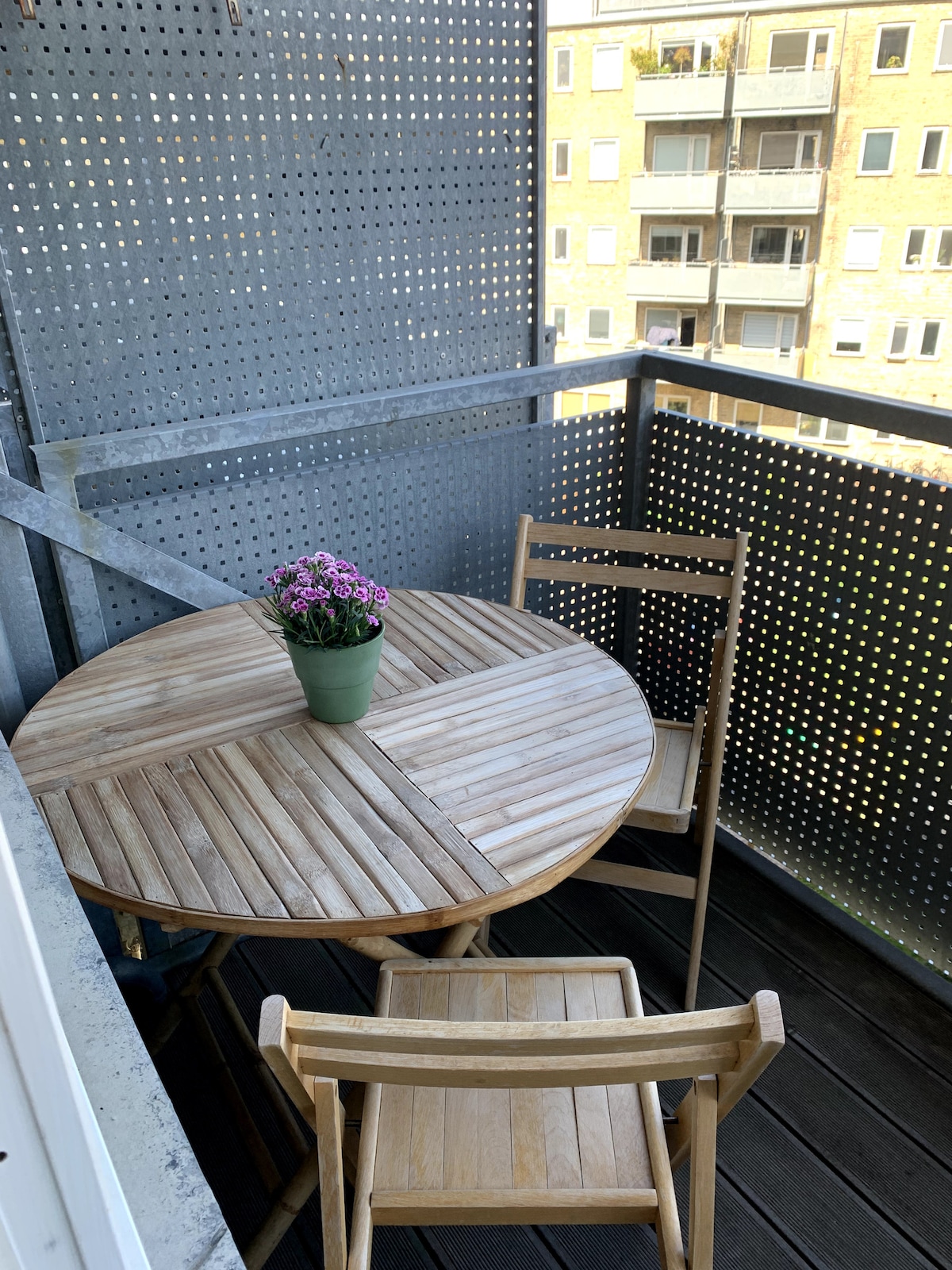 An outdoor balcony features a round wooden table surrounded by two folding chairs. A potted plant with purple flowers is centered on the table. The balcony's railing is adorned with a perforated design, allowing light and air to flow through.