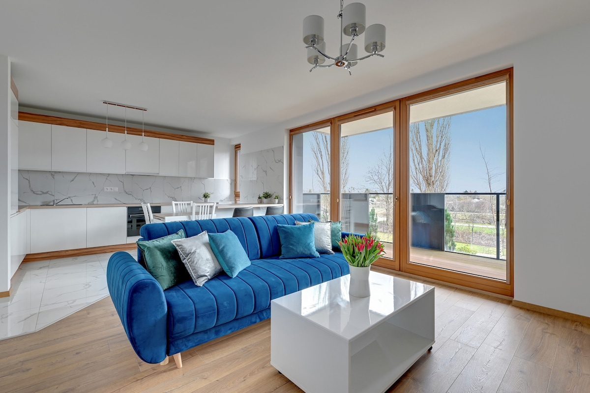 A spacious living area is furnished with a blue sofa adorned with cushions and a sleek white coffee table. Bright natural light filters through large windows with views of greenery outside, complementing the modern kitchen area in the background.