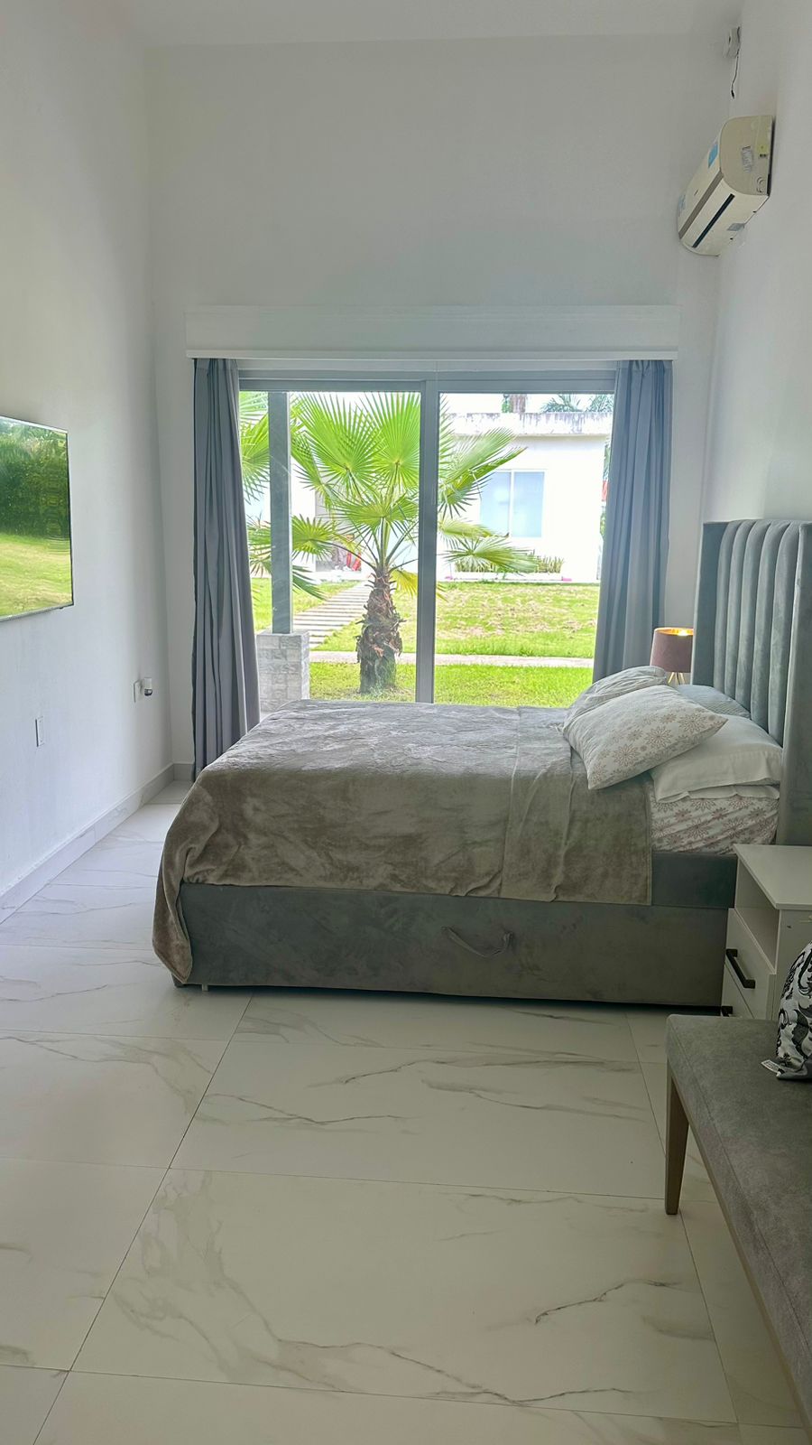 A serene sleeping area is illuminated by natural light from expansive glass doors leading to an outdoor view. A spacious bed is dressed in a soft gray coverlet, paired with decorative pillows. A comfortable armchair stands beside a bedside table, creating a cozy atmosphere.