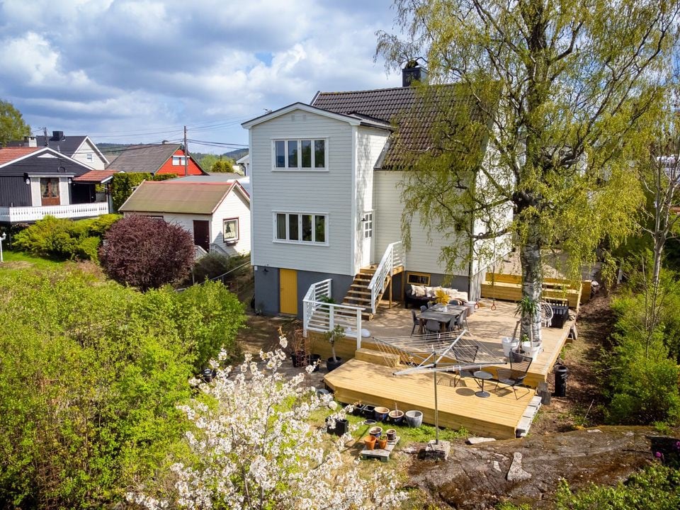 The exterior of the two-story house is surrounded by greenery, featuring a spacious wooden terrace with seating. A large tree provides shade over the area, while flowering plants add natural beauty. Nearby structures and landscapes create a serene neighborhood setting.