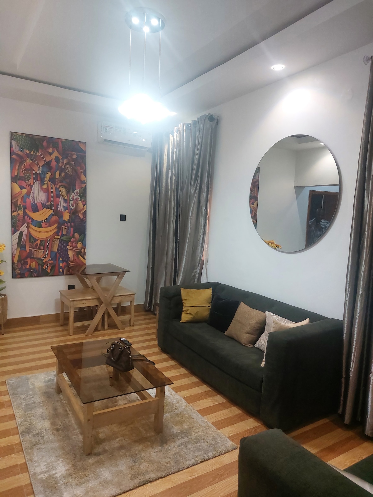A well-furnished living area features a cozy green sofa and matching armchair, complemented by a glass-top coffee table. A round mirror is hung on the wall opposite a vibrant piece of art. Light streaming from a ceiling fixture enhances the warm tones of the wooden floor.
