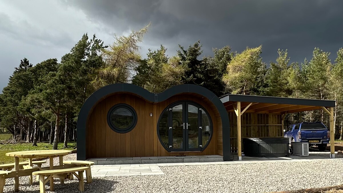 The lodge's exterior showcases a blend of wood and sleek design elements, featuring large circular windows that invite natural light. Surrounding greenery complements the structure, with a spacious patio area that includes a covered section and an outdoor hot tub.