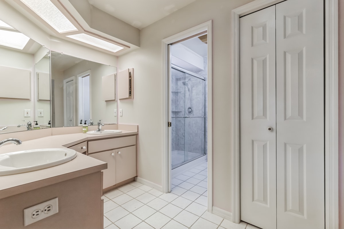 A spacious bathroom features a large vanity with dual sinks and ample counter space. The tiled floor adds to the practicality, while a door leads to a shower area and another door opens to storage. Natural light is provided by a frosted skylight.