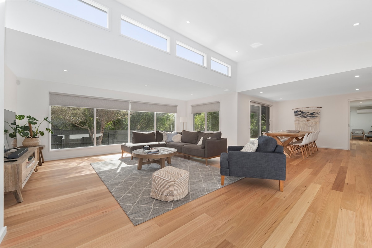 An open and airy living space features large windows allowing natural light to fill the room. A comfortable seating arrangement is complemented by a round coffee table on a textured area rug. A dining table is visible in the background, enhancing the spacious layout.