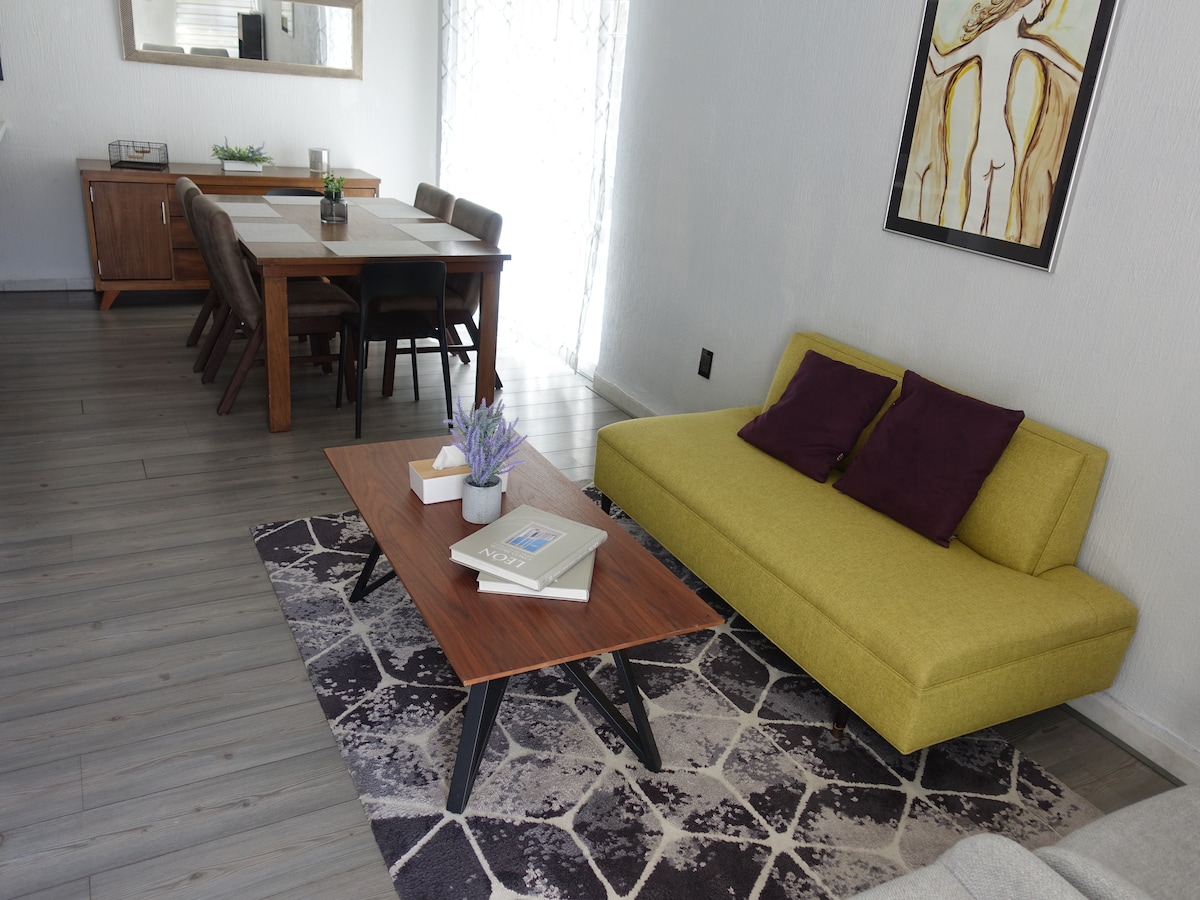 A modern living space is presented with a yellow sofa featuring purple cushions, paired with a wooden coffee table. A dining area with a table for six is visible in the background, along with a sideboard and a decorative mirror, providing a clean and inviting atmosphere.