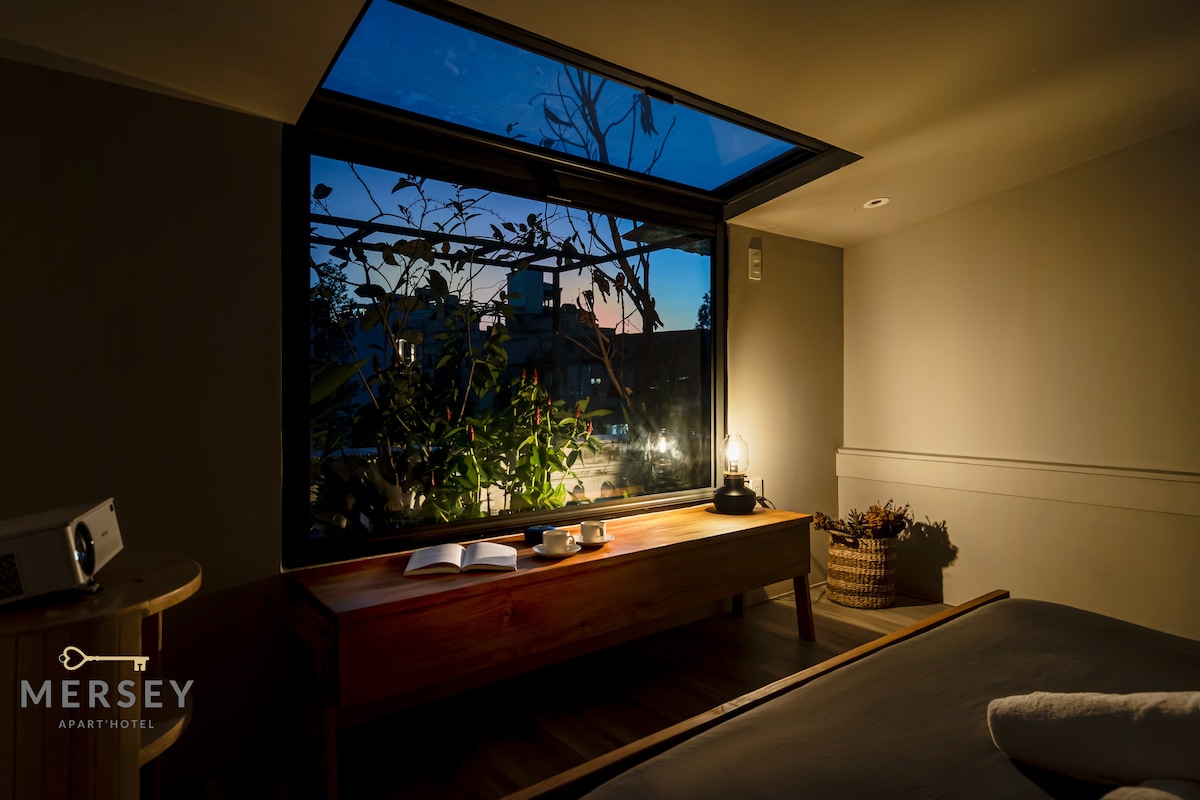 A modern room is adorned with a large window that allows natural light to filter in. A wooden bench sits beside the window, accompanied by a small lantern and an open book. Soft evening hues are visible beyond the glass, enhancing the peaceful ambiance.