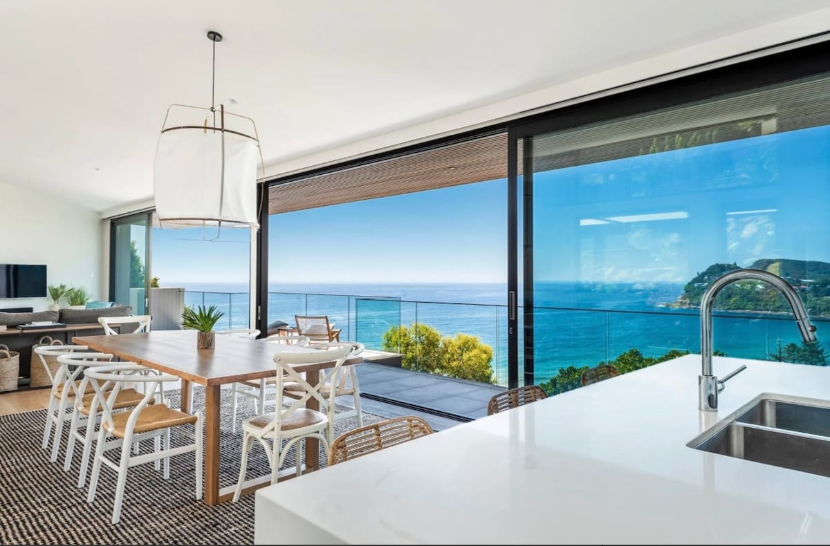 An open-plan dining and kitchen area features a wooden table surrounded by chairs, with expansive glass doors showcasing sweeping ocean views. A modern sink is positioned on a white countertop, and a softly glowing light fixture hangs above the table, creating an inviting space.