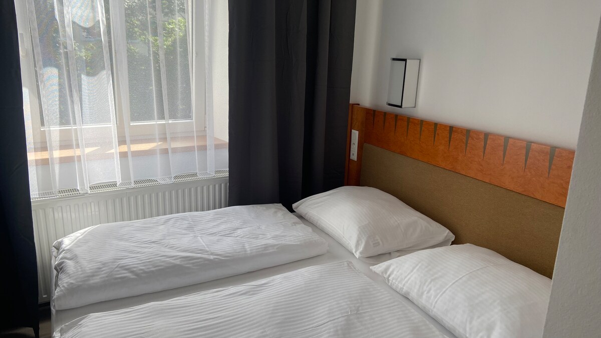A well-appointed sleeping area is featured, showcasing a spacious bed with light bedding. Sunlight filters through a large window, with sheer curtains providing a soft glow. Dark curtains frame the window, while a minimalistic wall sconce adds subtle illumination.