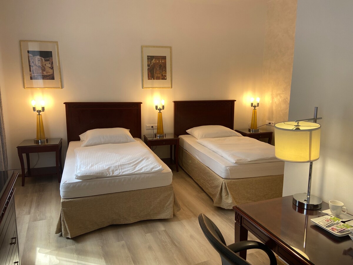 The spacious guest room features two individual beds with light bedding, positioned beside wooden nightstands with lamps. A desk and chair are visible, along with light-colored flooring and subtle wall decor, creating a clean and open atmosphere.