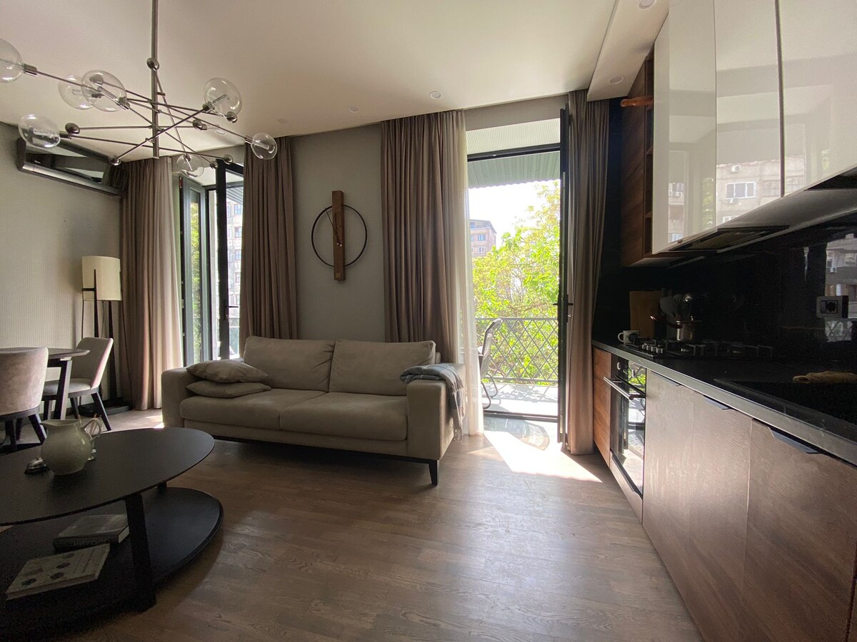 A cozy living space features a light-colored sofa positioned near large glass doors that open to a balcony. The room is designed with wood flooring and includes a dark, modern kitchen area with cabinetry and appliances visible. Natural light illuminates the space.