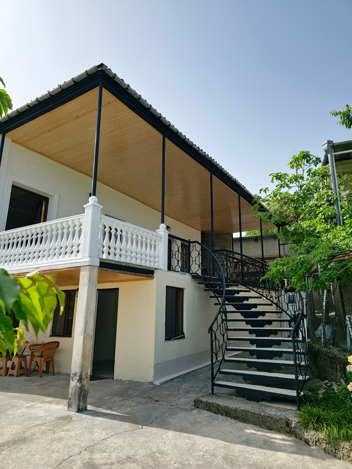 A two-story house is shown, featuring a covered balcony with decorative railings. A spiral staircase leads to the upper level, while two windows provide natural light to the lower level. Green foliage surrounds the entrance, enhancing the home's inviting exterior.