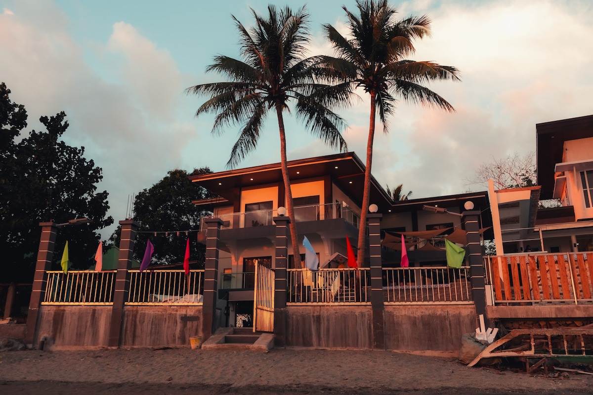 A beachfront property is framed by palm trees, featuring a wide porch with vibrant flags fluttering in the breeze. The modern facade is characterized by large windows and a contemporary design, creating an inviting entrance to the space.
