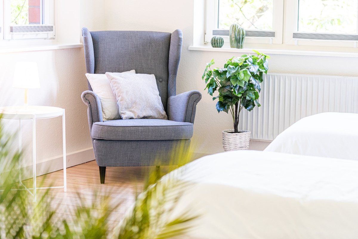 A cozy corner features a gray armchair adorned with soft cushions, positioned near the window. A small side table stands beside the chair, while a decorative plant in a white pot adds a touch of greenery to the inviting space.