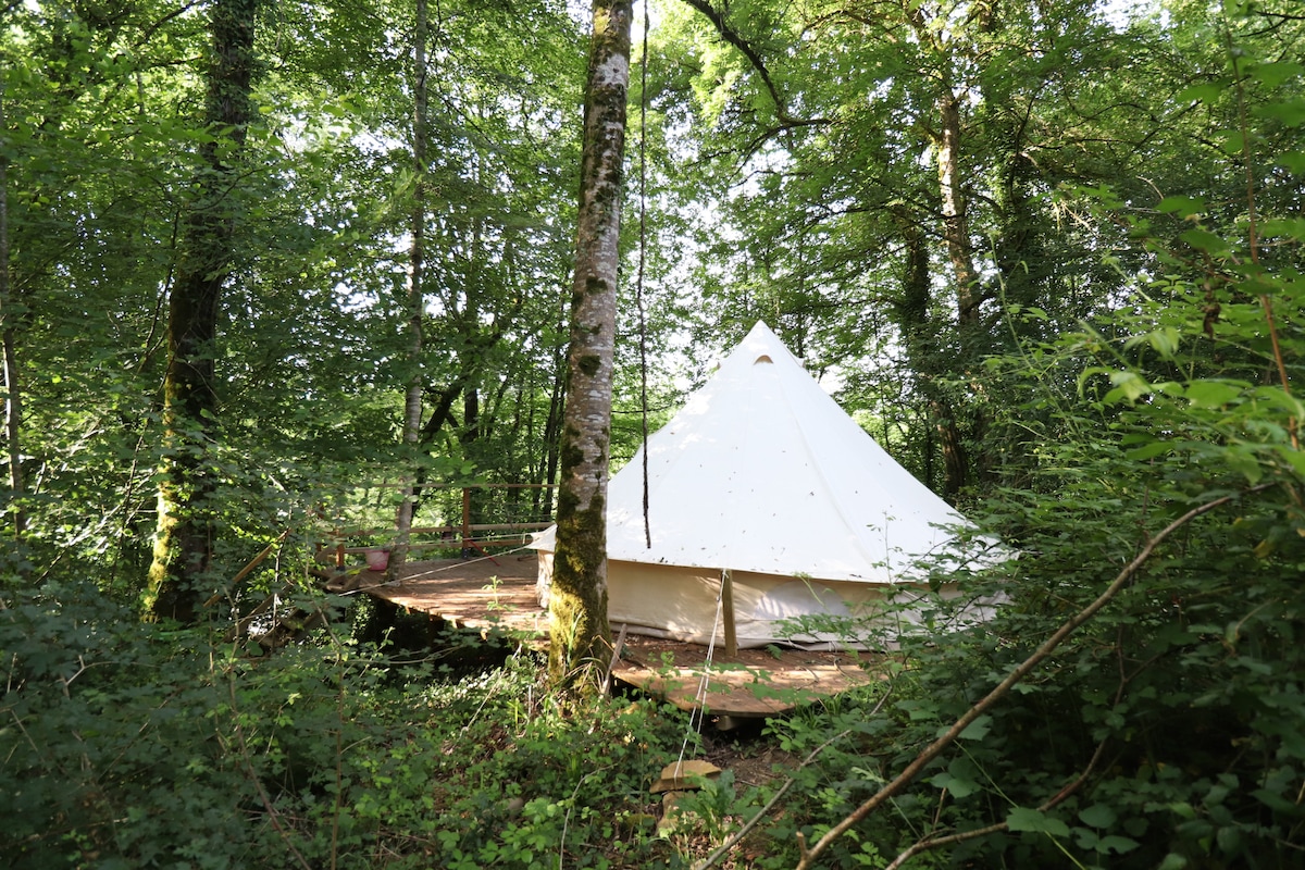 A spacious tipi tent is nestled among lush greenery, surrounded by tall trees. A wooden deck extends from the tent, creating a natural, serene environment. The white fabric of the tent contrasts with the vibrant green foliage, contributing to a tranquil outdoor setting.