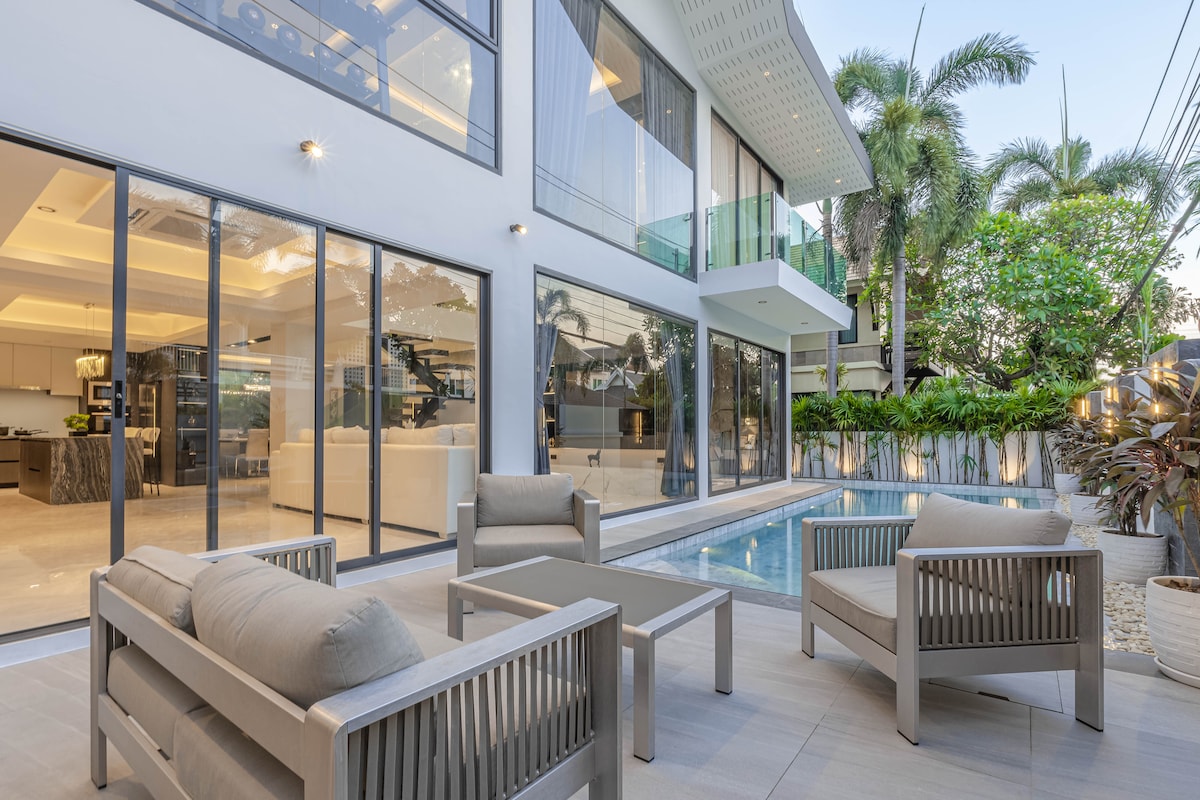 An outdoor seating area features two armchairs and a coffee table, arranged beside a private pool. The modern villa's expansive glass walls open to a bright living space, with lush greenery surrounding the property, creating a serene environment.