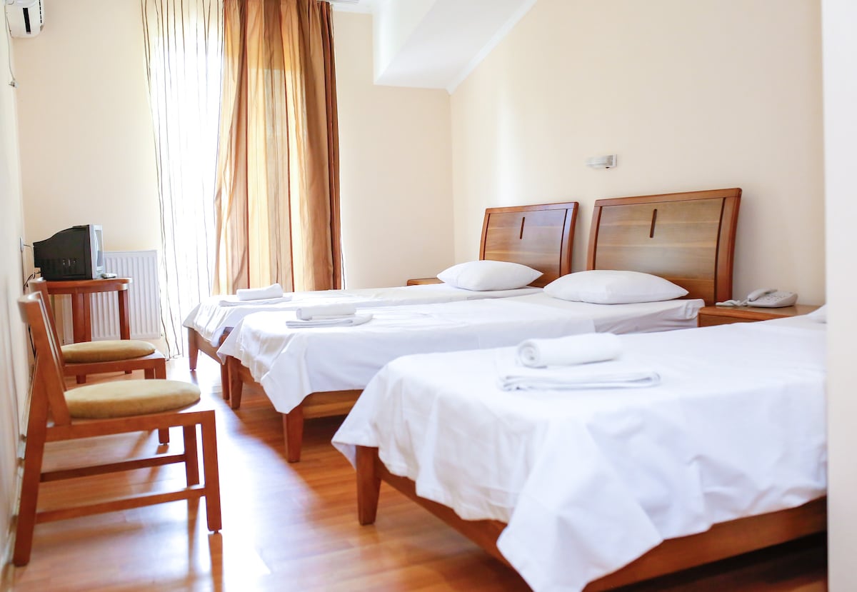 A well-lit room features two wooden beds with white linens, complemented by matching wooden chairs. Natural light enters through a large window adorned with sheer curtains, enhancing the calming ambiance. A small desk is positioned against the wall, adding functionality to the space.