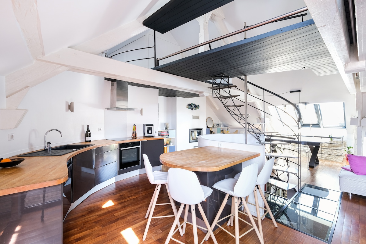 An open-concept kitchen and dining area featuring a round wooden table with white chairs. A black spiral staircase leads to the upper level. Large windows provide ample natural light, highlighting modern furnishings and wooden flooring throughout the space.