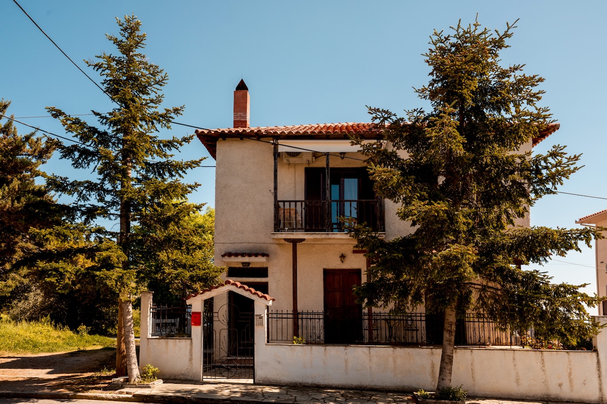 The exterior showcases a two-story house surrounded by tall trees, providing shade and a sense of privacy. A red door and balcony are prominent features, while a stone pathway leads to the entrance. Clear blue skies create a bright and pleasant atmosphere.