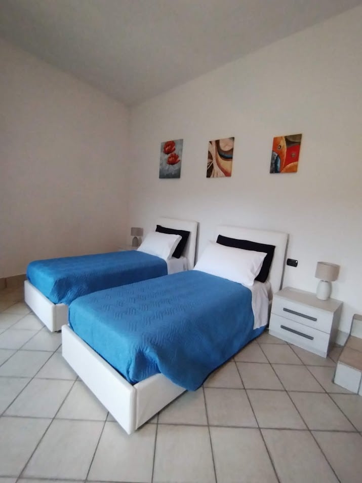 The bedroom features two twin beds covered in bright blue blankets. A bedside table with a lamp is positioned next to each bed. Abstract artwork adorns the wall above the beds, along with ample natural light illuminating the neutral-toned space.
