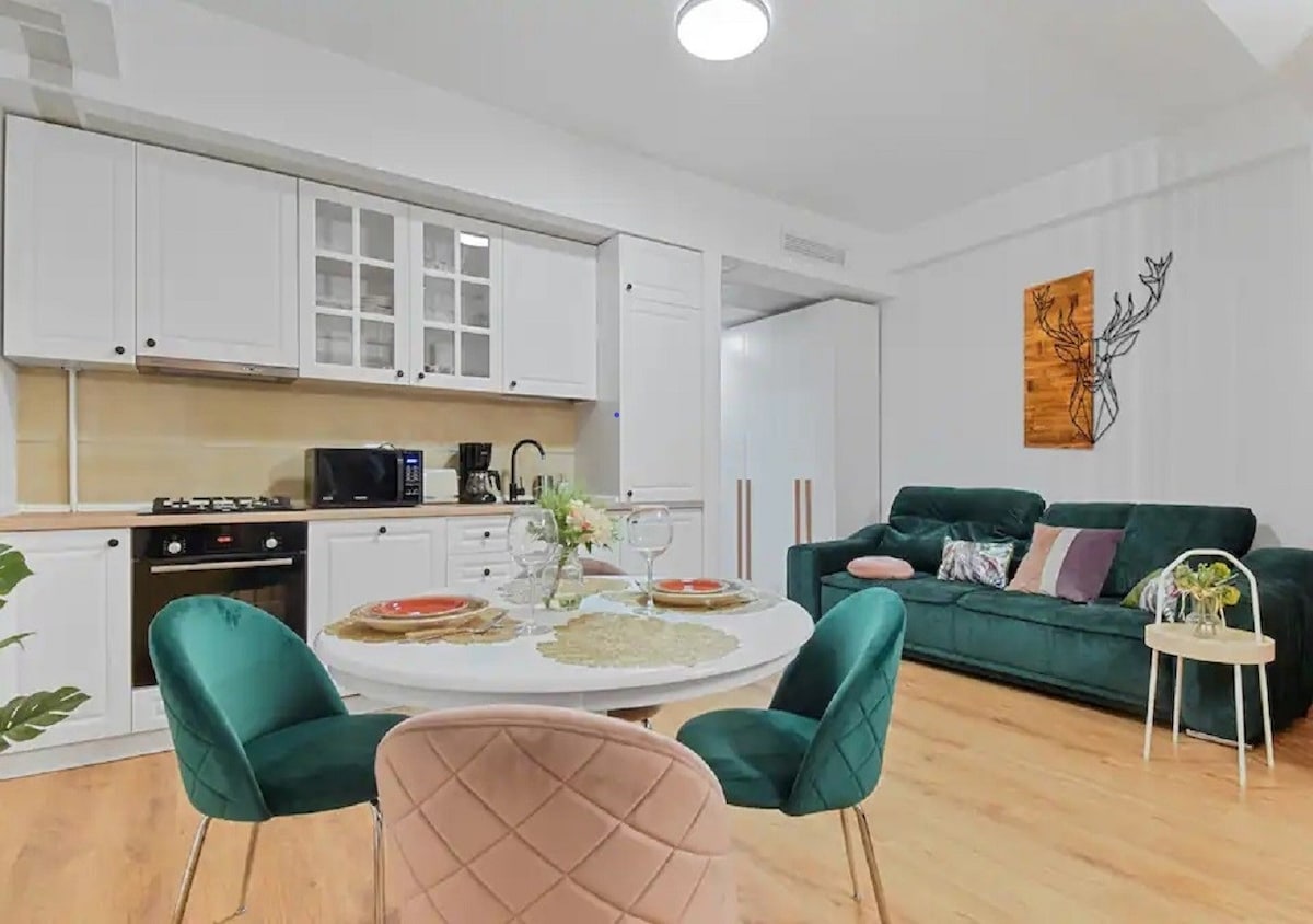 A modern kitchen area features white cabinetry and stainless-steel appliances, complemented by a round dining table set for two. A cozy green sofa is positioned nearby, adorned with colorful pillows, providing a welcoming space for relaxation.
