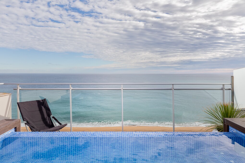 A private pool is visible, featuring blue tiles that reflect the sky. The view beyond includes a calm sea and an expansive shoreline under a partly cloudy sky. A black lounge chair is positioned nearby, offering a place to relax and enjoy the scenery.