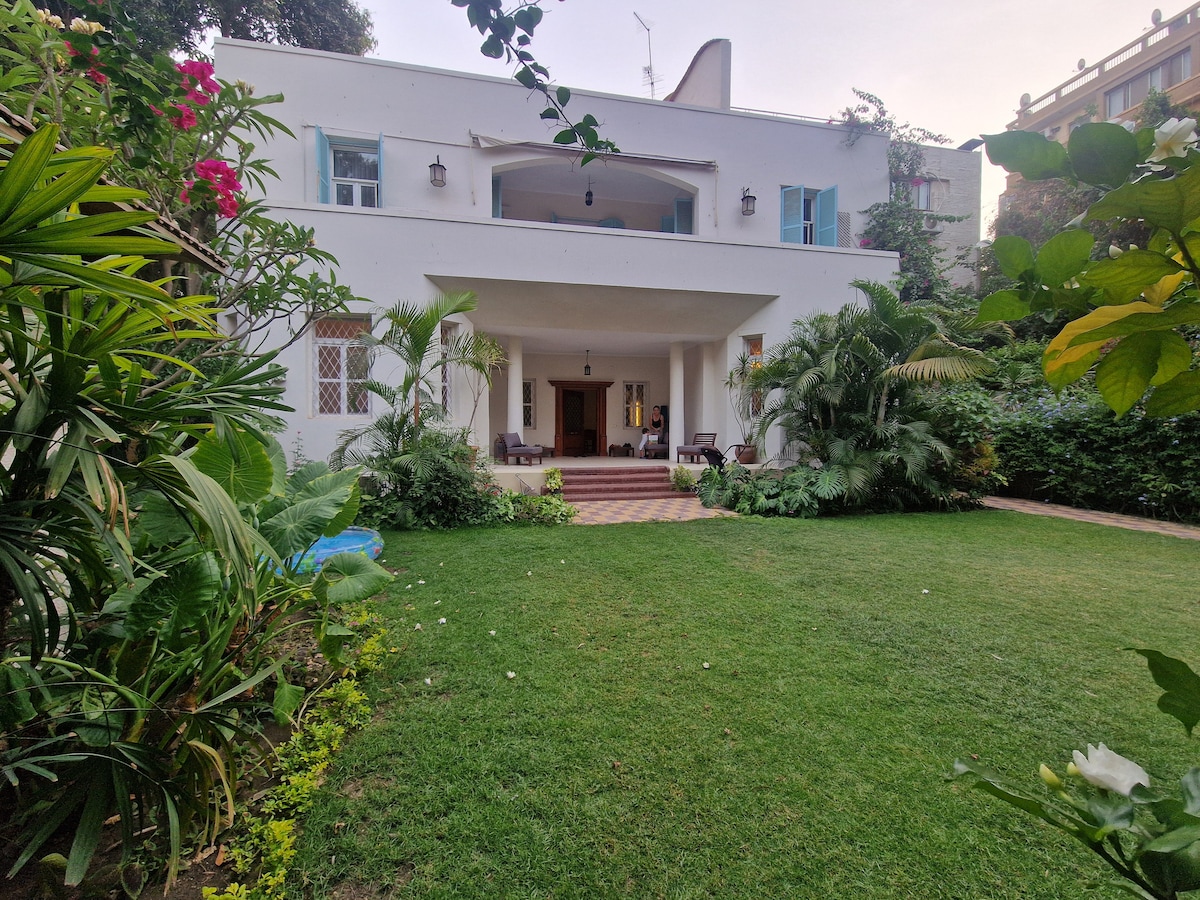 The villa is framed by lush greenery in a well-maintained garden. The spacious entrance features a covered porch with two dark wooden chairs. A pathway leads to the main door, surrounded by vibrant flowering plants and shrubs that create a welcoming atmosphere.