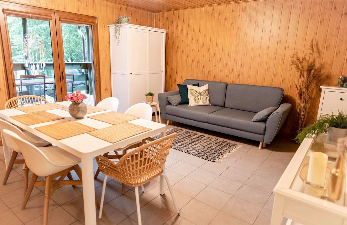 A cozy dining and living area features a white table surrounded by six chairs, complemented by a gray sofa. Natural wood paneling creates warmth, while large sliding doors provide a view of the outdoors. Subtle decorative elements enhance the space.