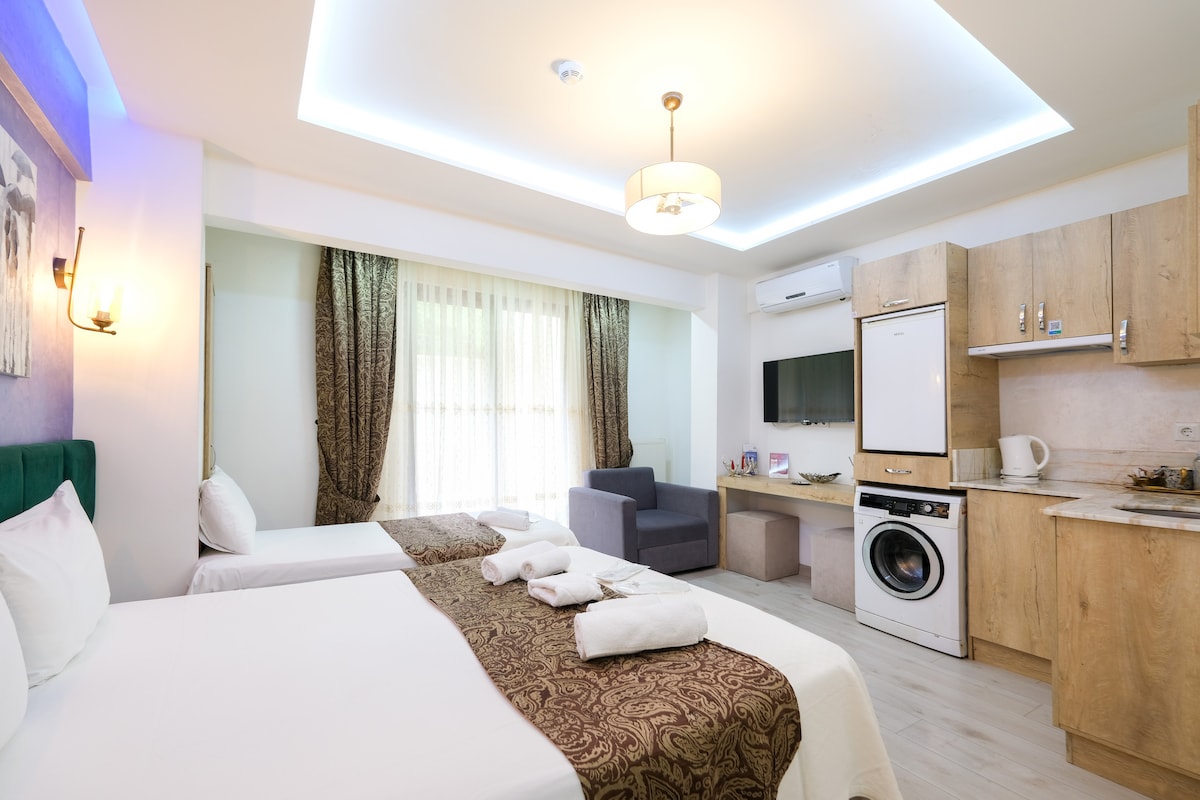 A room is designed with two beds, each featuring neatly arranged linens. A kitchenette equipped with essential appliances is visible. Large windows allow natural light to fill the space, with flowing curtains adding a soft touch. A seating area with a gray chair is also present.