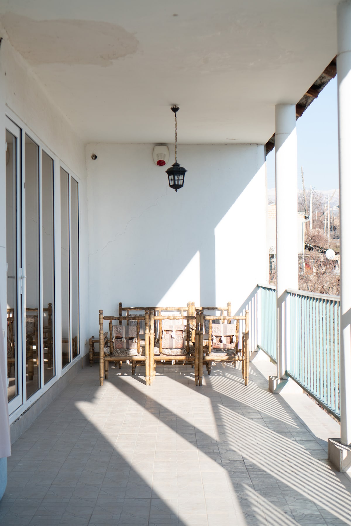 A covered terrace space is presented, featuring a set of bamboo chairs arranged around a small table. Large glass doors reflect light, while a lantern hangs from the ceiling, casting a gentle shadow across the tiled floor.
