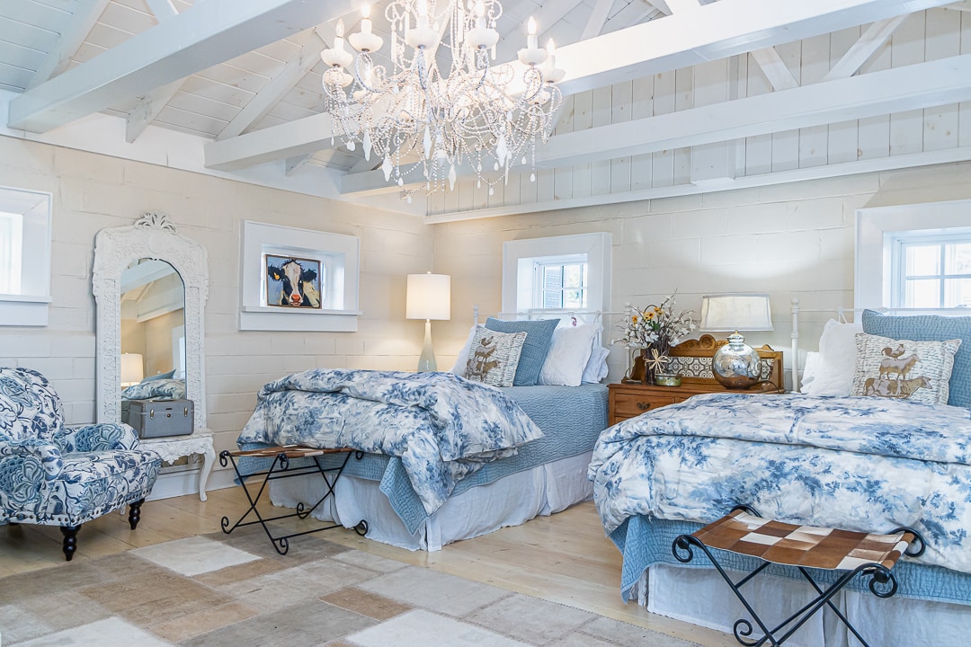 A bright bedroom features two spacious queen beds adorned with floral-patterned bedding. A modern chandelier hangs from the beamed ceiling, offering soft light. A decorative mirror is positioned next to a cozy chair, with a bedside table showcasing a decorative vase.