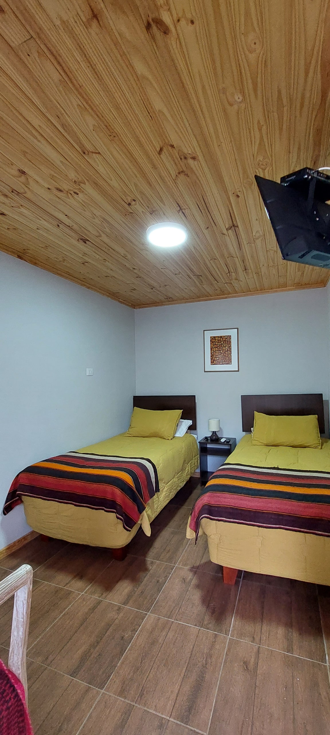 A bright bedroom is presented with two twin beds, each topped with yellow bedding and striped throws. A simple nightstand stands between the beds, supporting a small lamp. The walls are painted light gray, complemented by a warm wooden ceiling.