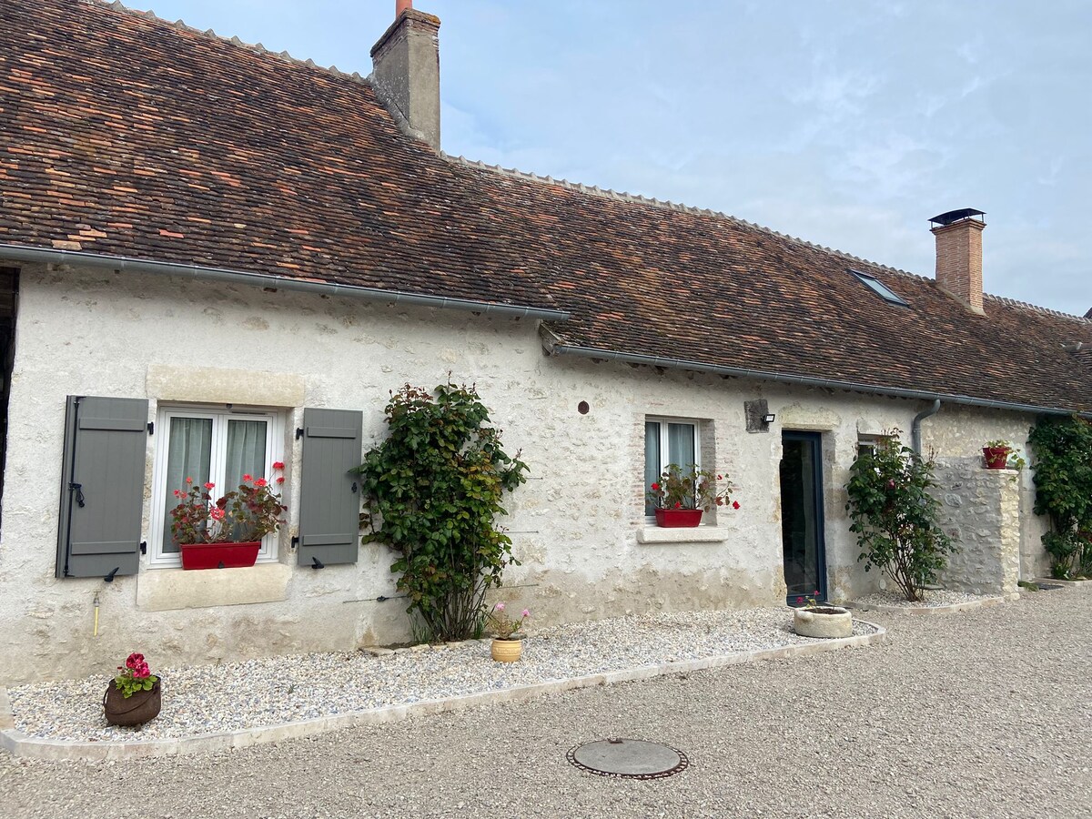 The exterior of the gîte is shown, featuring a traditional stone façade with wooden shutters. Flower boxes filled with vibrant blooms adorn the windows. The gravel driveway leads to the entrance, framed by lush greenery and planters, creating an inviting atmosphere.