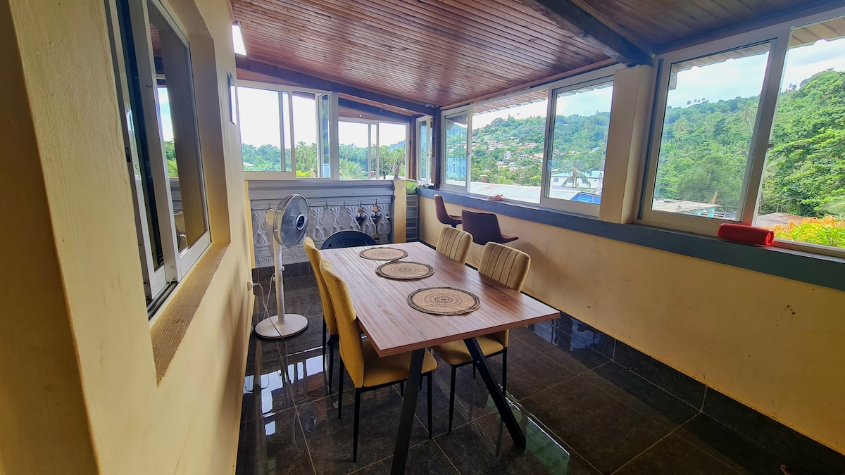 An airy dining area is presented, featuring a large wooden table surrounded by six upholstered chairs. Floor-to-ceiling windows offer views of the greenery outside. A standing fan is positioned in the corner, contributing to the room's comfort and openness.