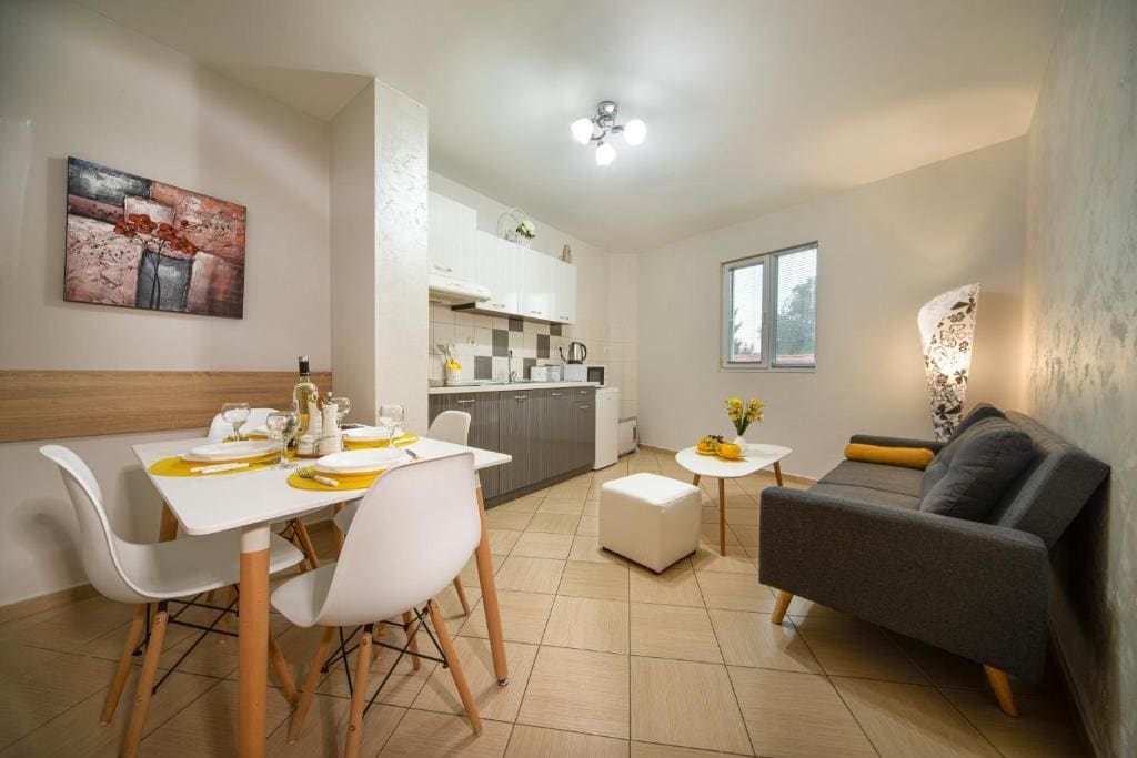 A bright and modern dining area features a white table set for four, surrounded by sleek chairs. Light-colored cabinetry and contrasting tiles are visible in the adjacent kitchen, while a cozy grey sofa and a small white ottoman create a comfortable lounge space.