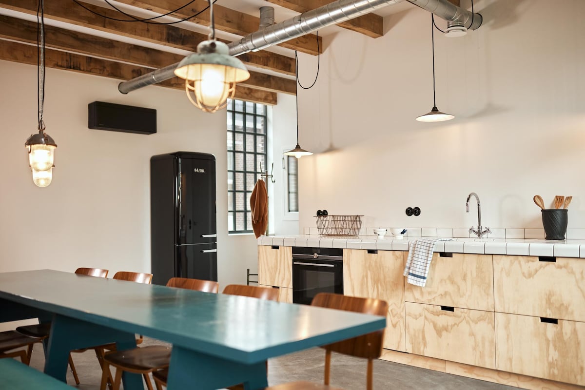 A spacious kitchen features a long table surrounded by wooden chairs. A modern black refrigerator stands nearby, while a minimalist countertop showcases essential kitchen tools. Industrial lighting hangs from the ceiling beams, illuminating the room's clean lines and functional design.