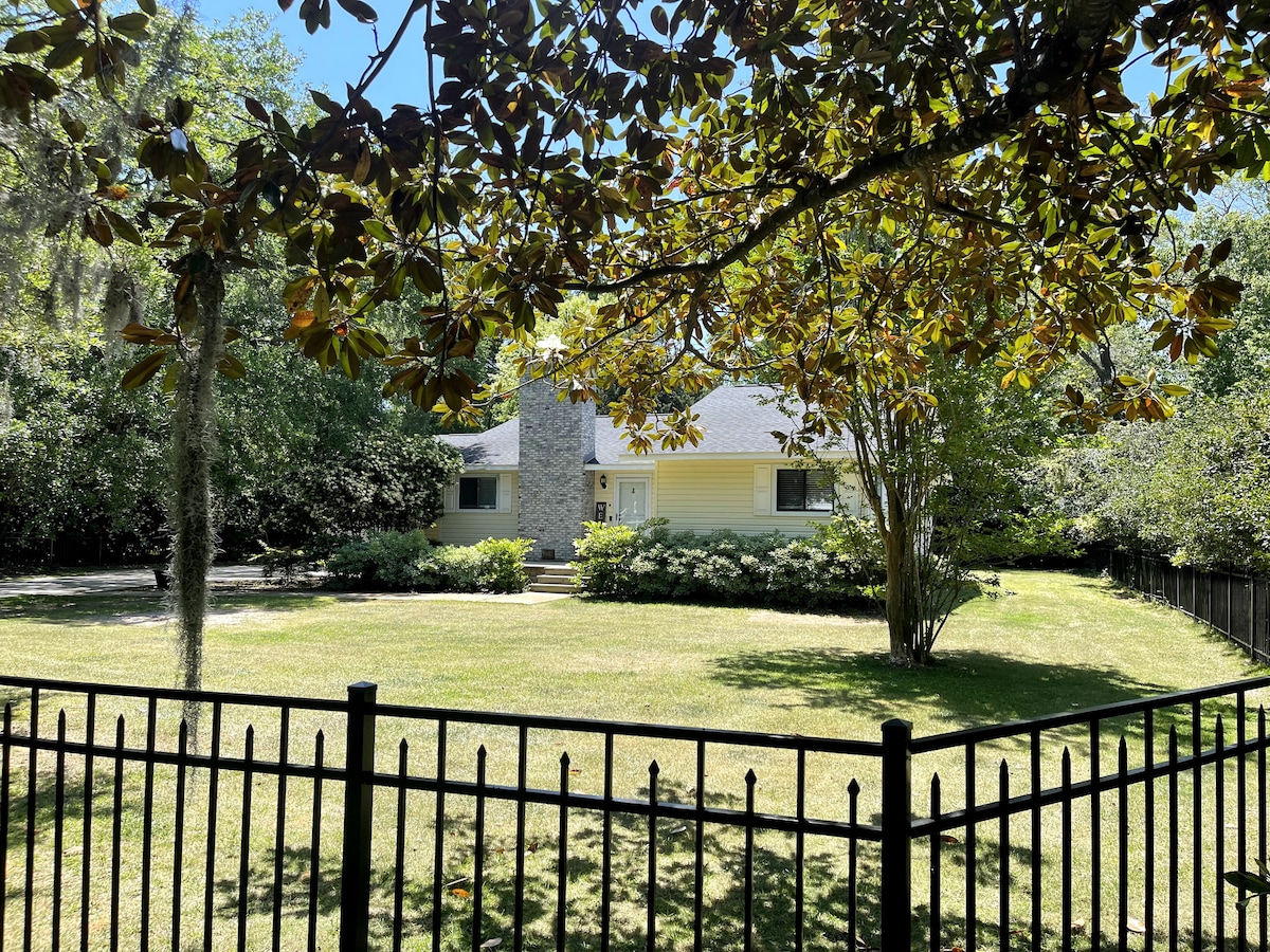 A charming house is framed by lush greenery, featuring a stone accent on the front. A spacious, grassy yard is enclosed by a black, wrought iron fence, with mature trees providing shade and a peaceful ambiance.