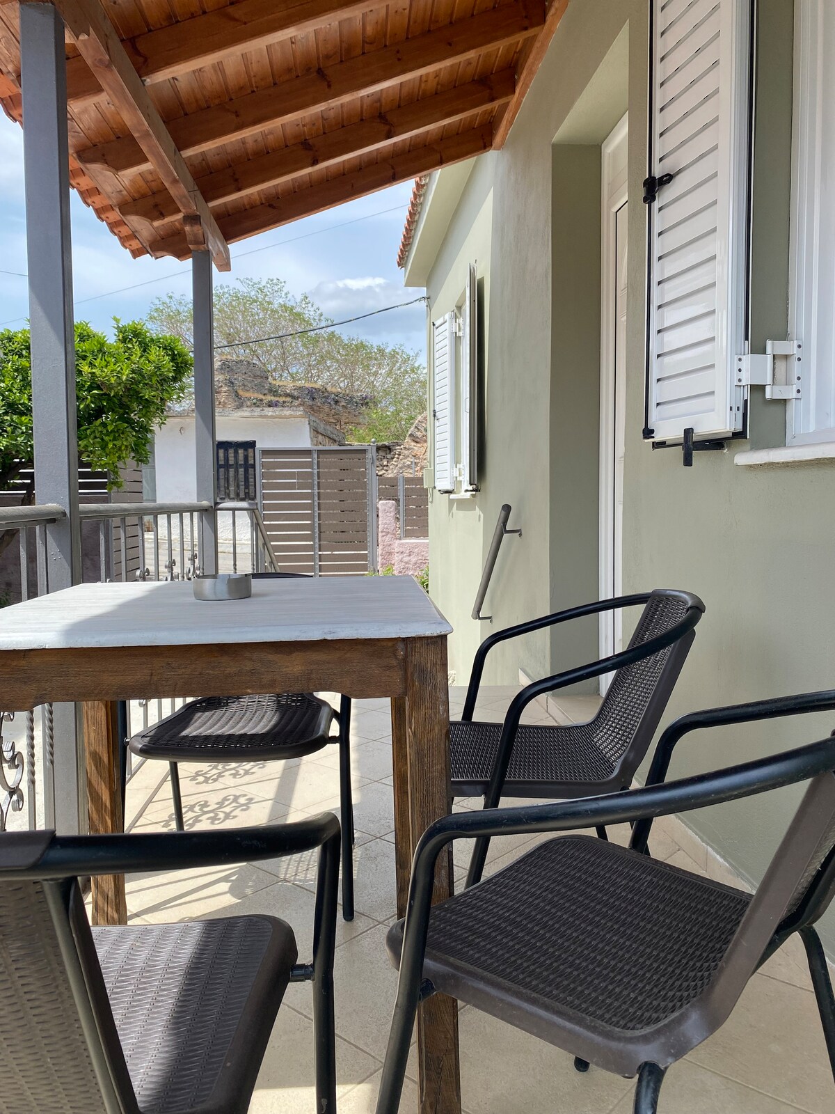 An outdoor seating area is presented with a wooden table and four black chairs. Lush greenery is visible in the background, along with a shaded space created by a wooden roof. Light streams in, highlighting the inviting outdoor setting.