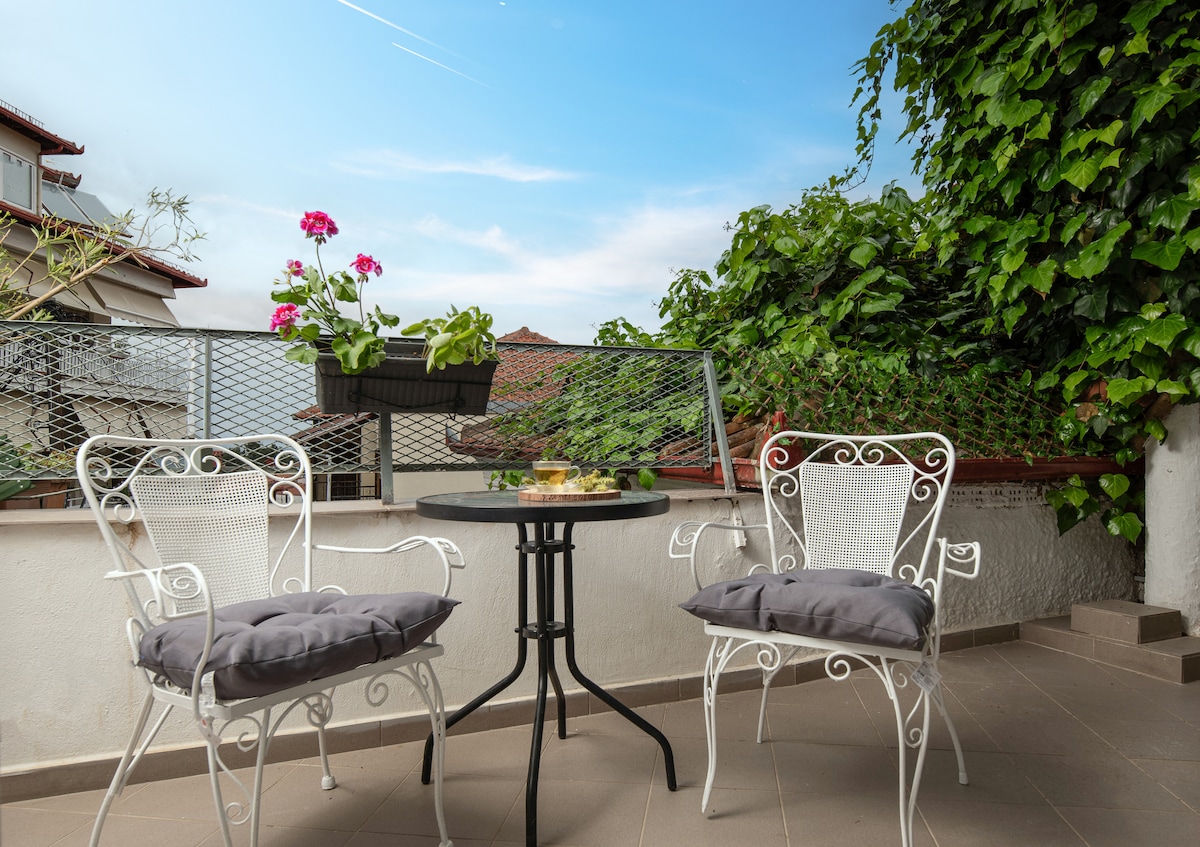 A small outdoor terrace features two white wrought iron chairs with gray cushions positioned around a round table. Potted flowers add a touch of color, while lush greenery provides privacy and a natural backdrop. The sky above is bright and clear, creating a serene atmosphere.
