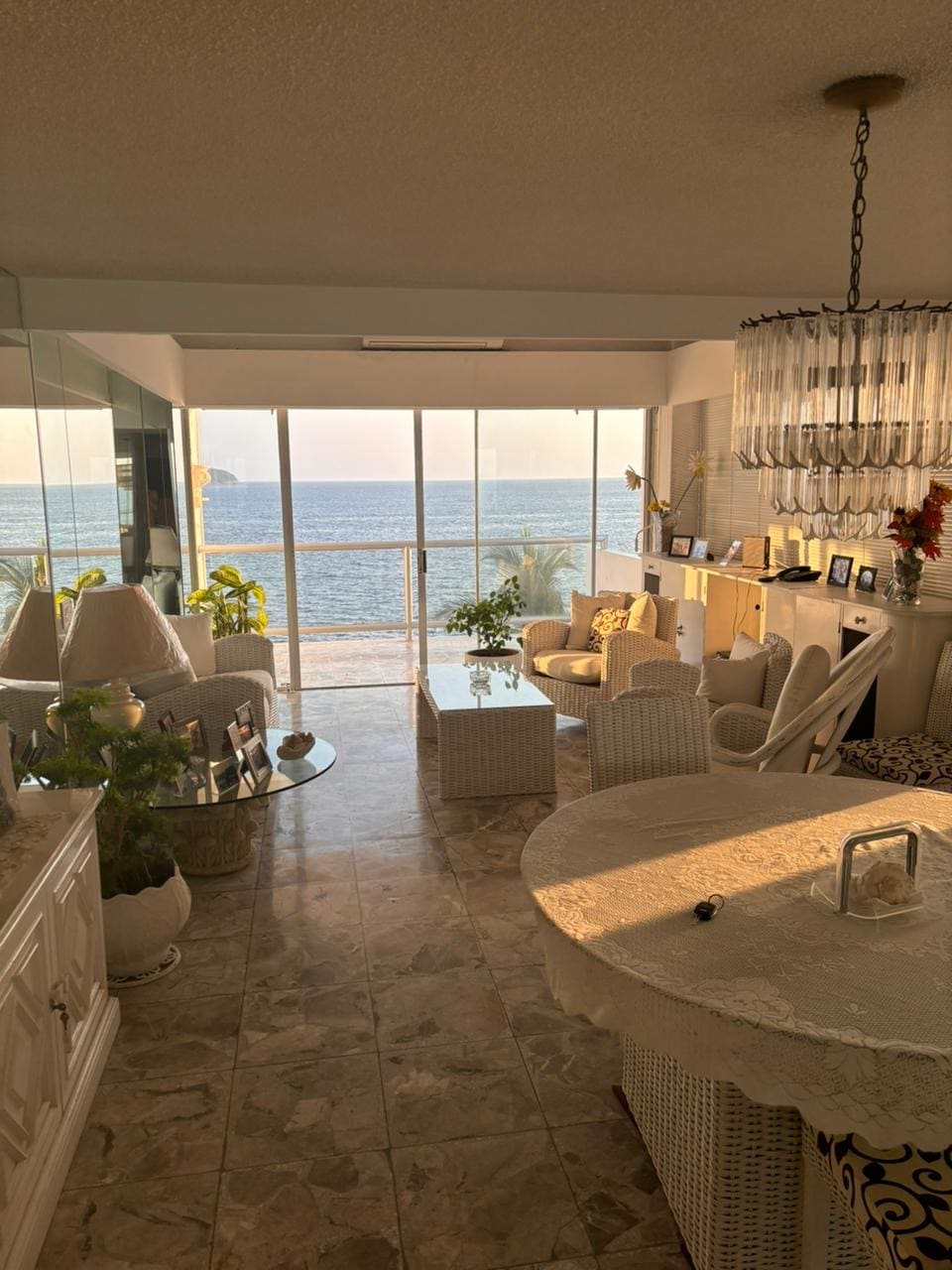 A spacious living area features large glass windows overlooking the ocean, allowing natural light to fill the room. Stylish white furniture complements the serene atmosphere. A round dining table is positioned near the window, surrounded by tasteful decor and greenery.