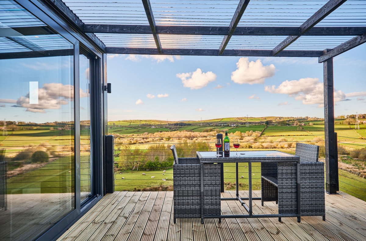 A covered outdoor patio features a stylish table set for two, accompanied by glasses of wine. The surrounding landscape showcases expansive green fields and distant hills, with a clear sky dotted by fluffy clouds, providing a serene backdrop for relaxation.