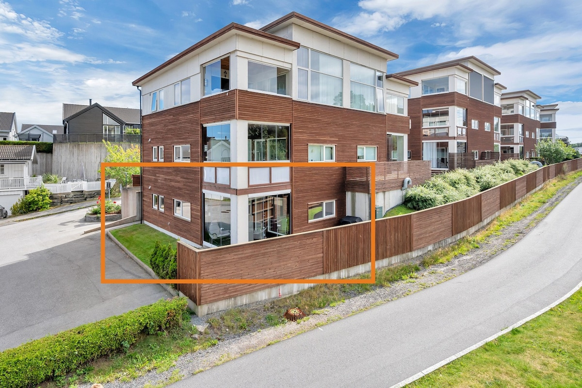 A modern building with large glass windows is shown, featuring multiple levels and a contemporary facade. Surrounding greenery and a paved pathway are visible, enhancing accessibility. The parking area is nearby, providing convenient access to the entrance.