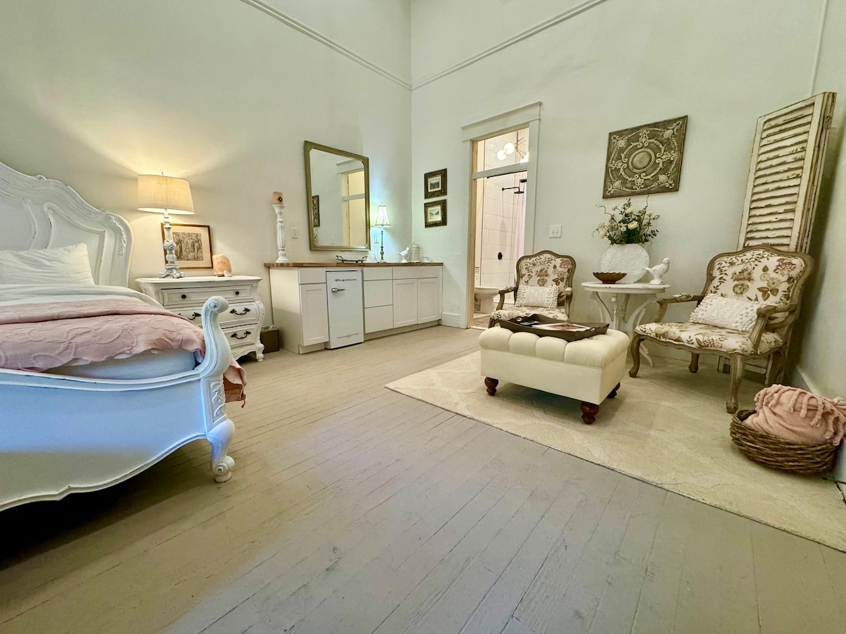 The spacious suite features a light-filled room with high ceilings. A large bed is complemented by a bedside lamp and decorative mirror. An elegant dresser and two cozy chairs are present, with a rug adding warmth to the wooden floor.
