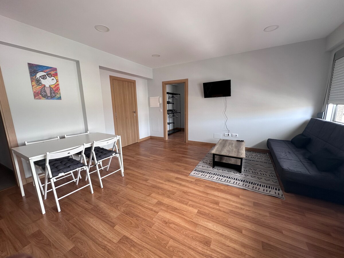 An open living area is shown with smooth wooden flooring. A stylish black sofa bed is positioned next to a wall-mounted television. A dining table with six white chairs is visible, complemented by a patterned area rug beneath. A doorway leads to another room.