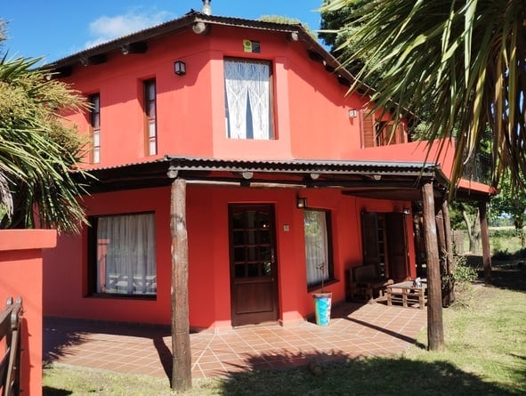 A two-story, red-painted cabin is showcased, featuring a covered porch supported by wooden beams. Large windows with white curtains allow natural light to enter. Surrounded by greenery, the outdoor space highlights a relaxed atmosphere in a tranquil setting.