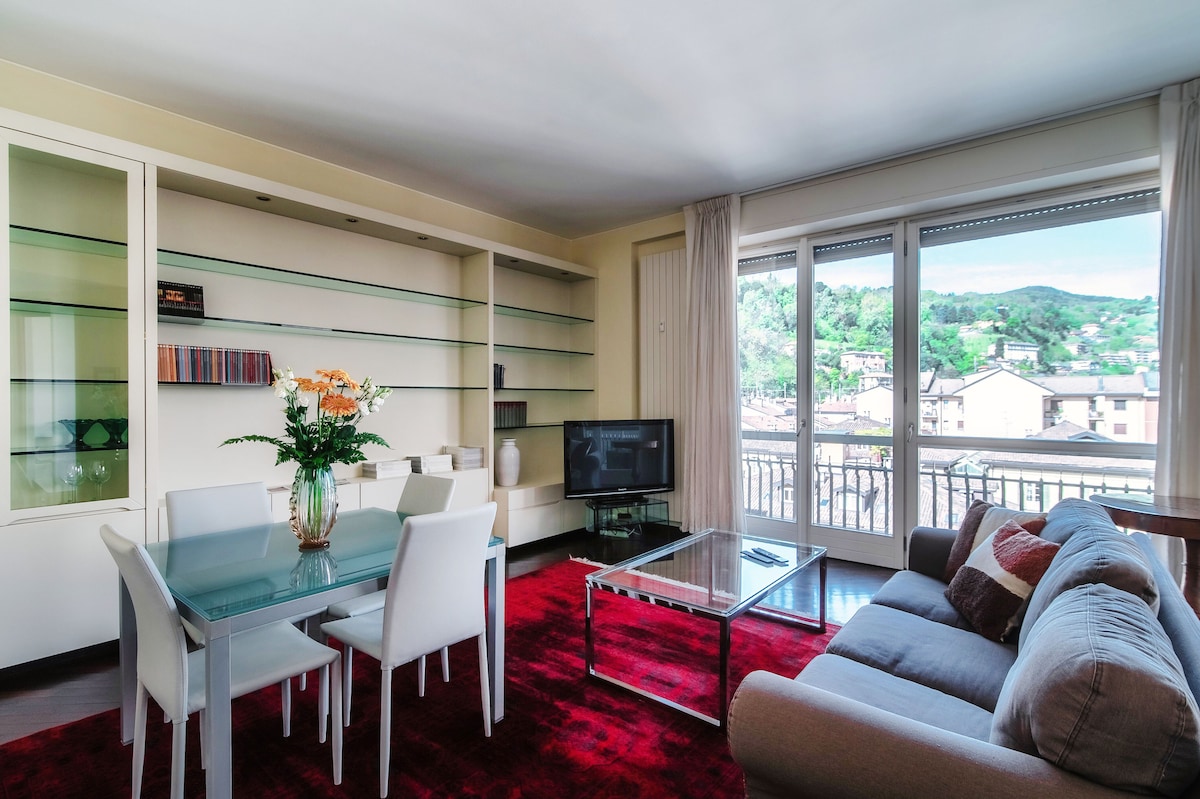 A bright living area features a glass dining table surrounded by white chairs, complemented by a cozy gray sofa. A glass coffee table sits on a red area rug, and a sleek TV is positioned against a modern shelving unit. Large windows offer views of the surrounding landscape.